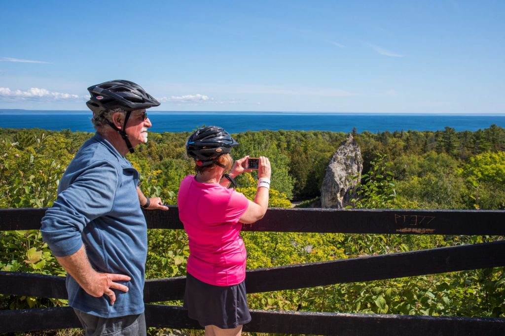 Man and Woman in Bicycle Helmets Taking Photo of Sugar Loaf Rock Formation in Distance on Mackinac Island