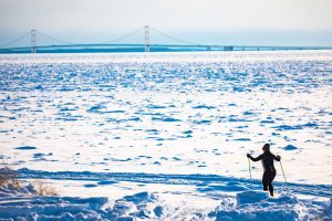 A cross-country skier traverses the snowy landscape along the shore of Mackinac Island with the Mackinac Bridge in the background