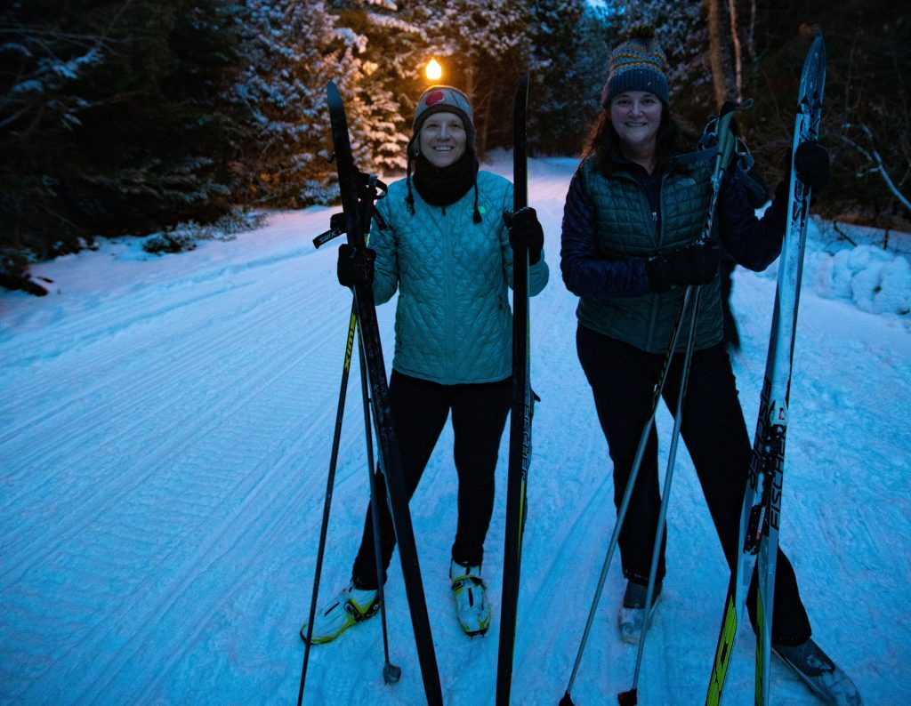 Two women stand with their cross-country skis and poles on a lantern-lit trail through Mackinac Island State Park