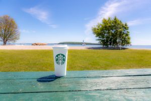 A cup of Starbucks coffee sits on a green picnic table at Mackinac Island's Windermere Point with water and lighthouse in background