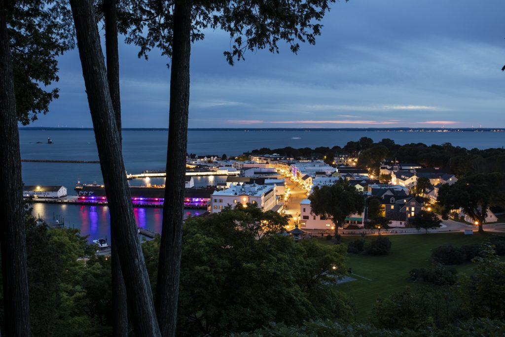 Downtown Mackinac Island is lit up at night as seen from above on the bluffs of Fort Mackinac overlooking the harbor.