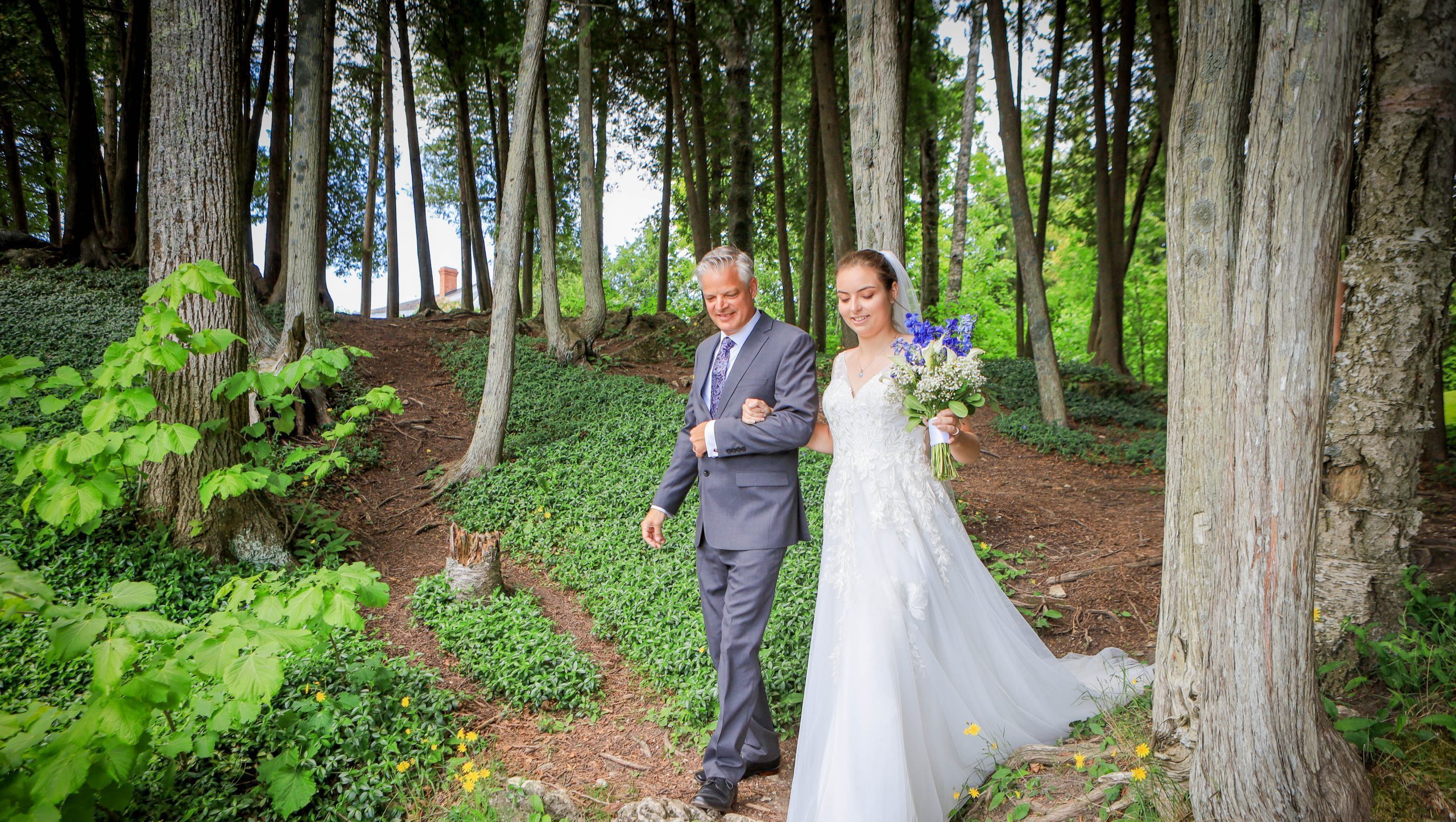 A bride and her father walk down the aisle through the woods at an outdoor wedding ceremony on Mackinac Island