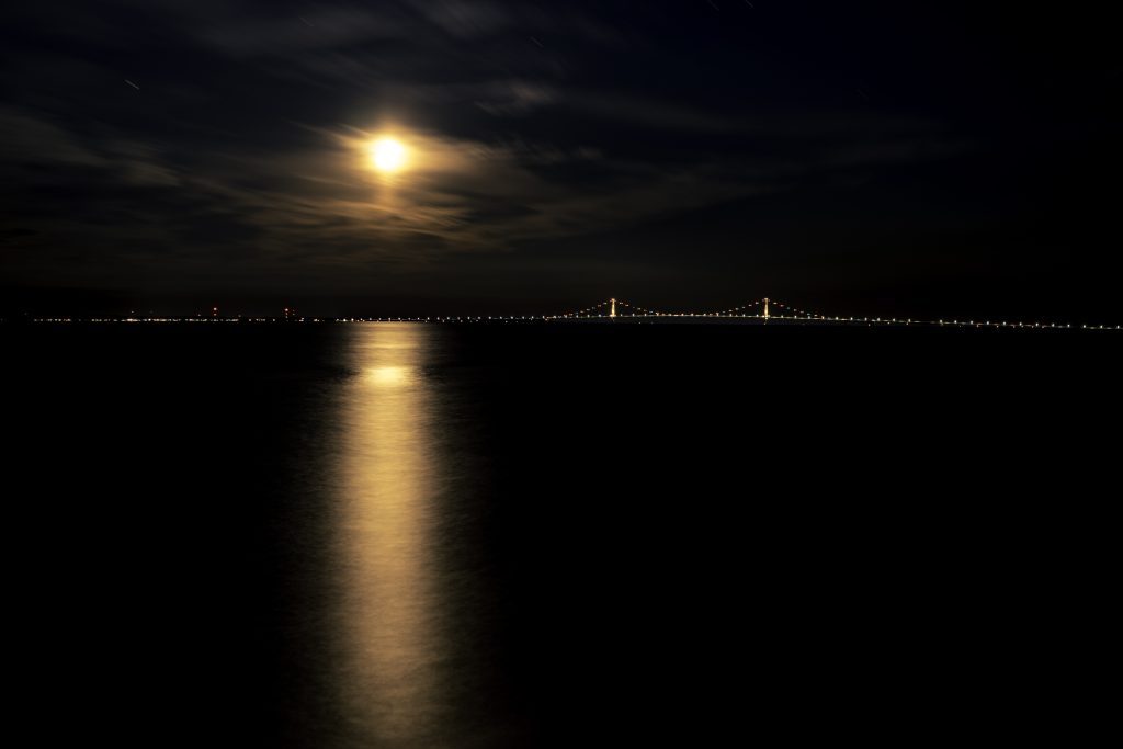 A full moon shines through the clouds above the water off Mackinac Island and a lighted Mackinac Bridge