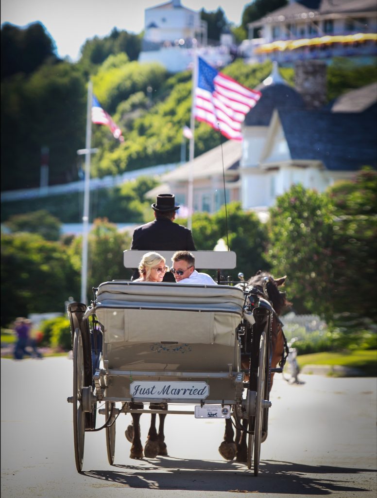 A bride and groom look back at the camera from the seat of a horse-drawn carriage going down the street in front of Fort Mackinac