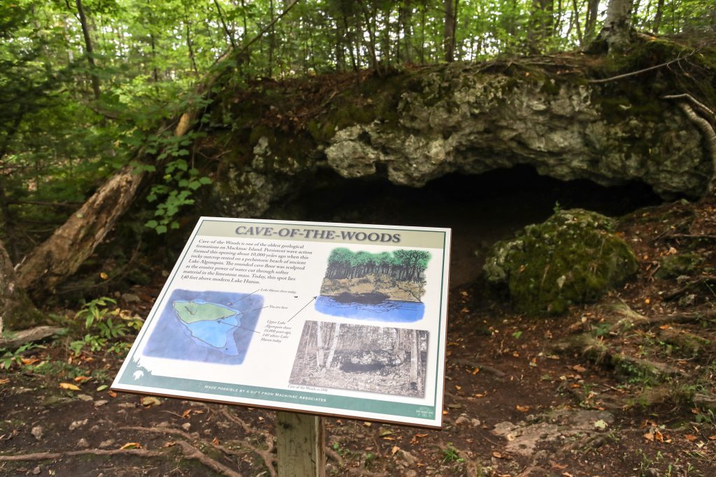 A sign for Cave of the Woods in Mackinac Island State Park details the history of the cave behind it.