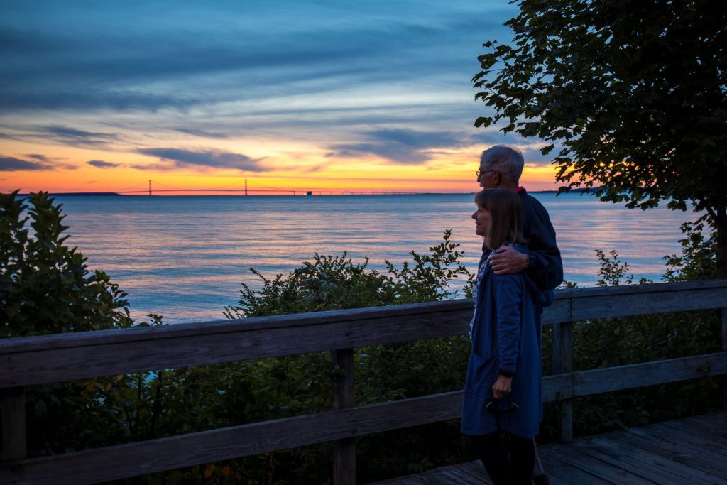 A couple walks hand in hand down the Mackinac Island boardwalk at sunset with the Mackinac Bridge in the background