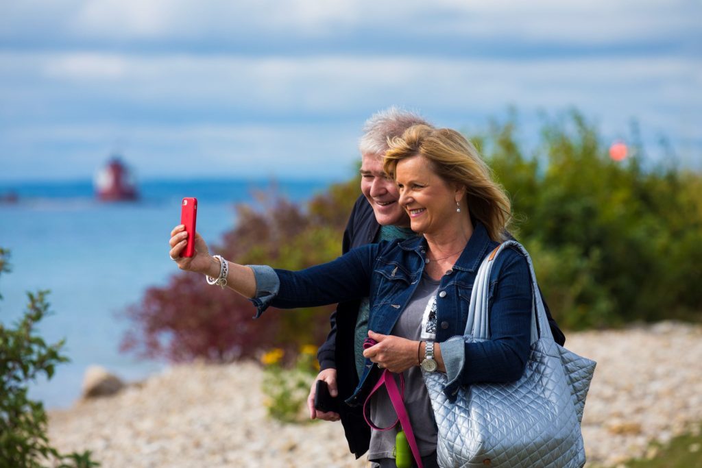 A couple poses for a selfie on a rocky Mackinac Island beach with Round Island Lighthouse in the background