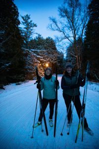 Two women smile for the camera while holding cross-country skis on a snowy Mackinac Island trail