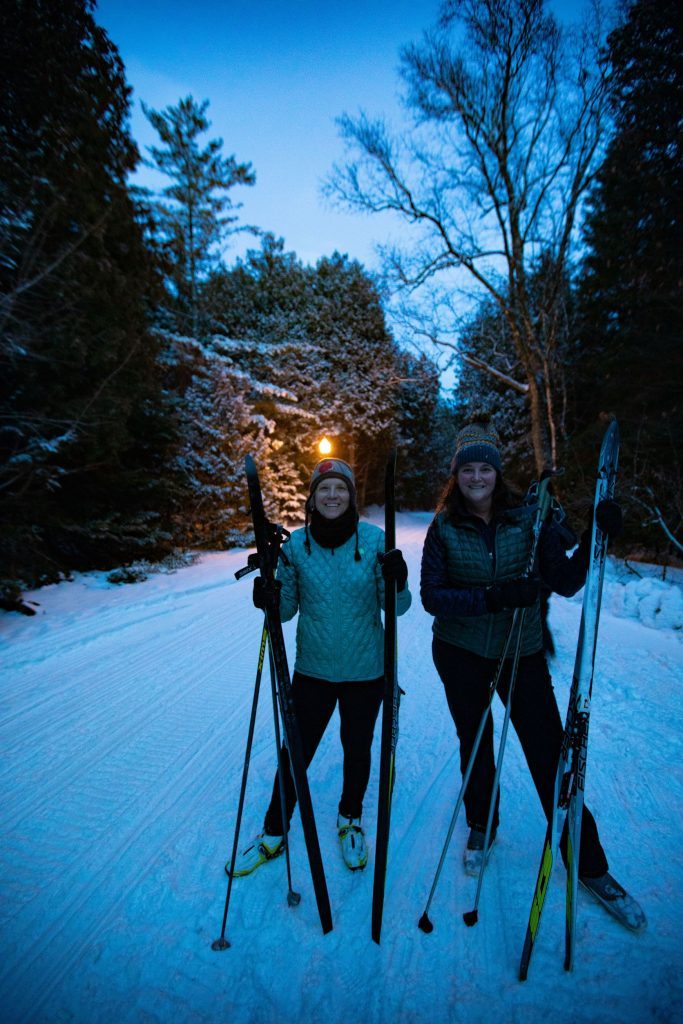 Two women smile for the camera while holding cross-country skis on a snowy Mackinac Island trail
