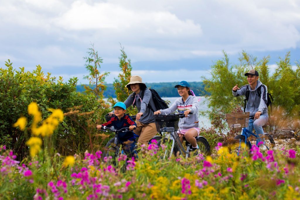 A family rides bikes on Mackinac Island along a waterfront trail lined with colorful flowers.