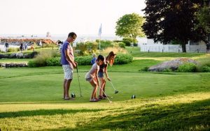 A father and two girls play putt putt along the water on Mackinac Island's Greens of Mackinac natural grass putting course