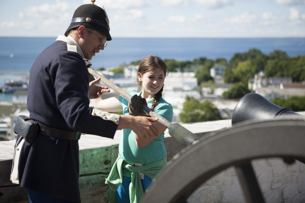 A historical interpreter at Fort Mackinac helps a teenage girl prepare to blast the cannon over Mackinac Island 