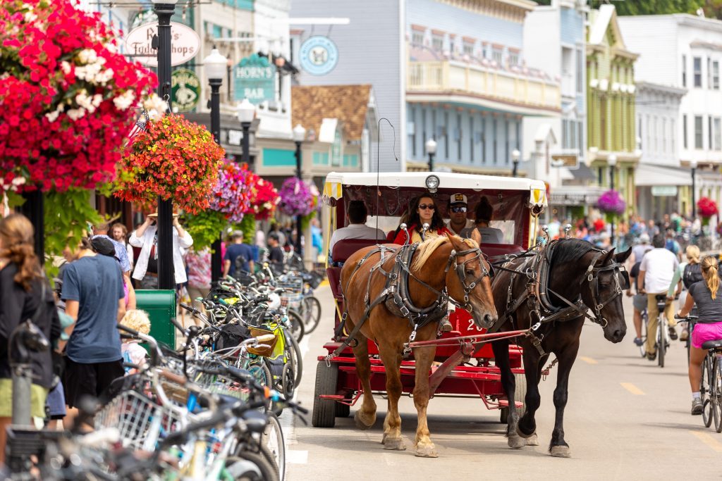 A horse-drawn carriage drives through downtown Mackinac Island with bicycles and flowers on the side of the street