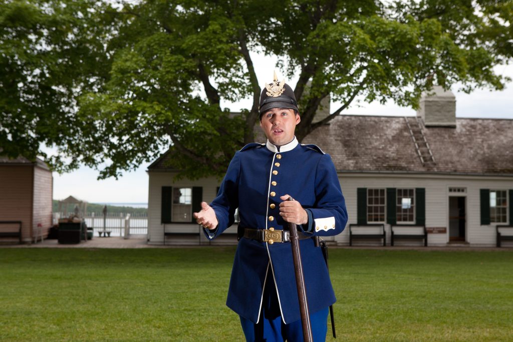 A historical interpreter at Fort Mackinac stands in the midst of the fort with an historic building in the background