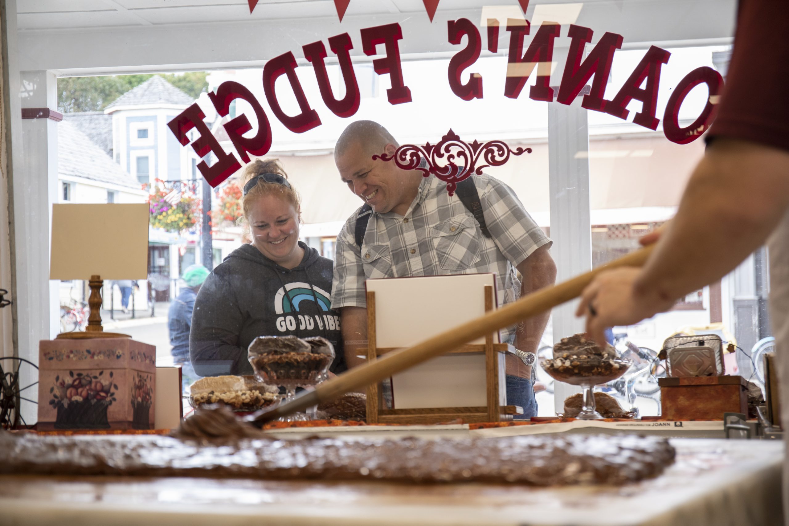 A couple peeks through the front window of a Mackinac Island fudge shop as a fudgemaker paddles gooey chocolate into a loaf.