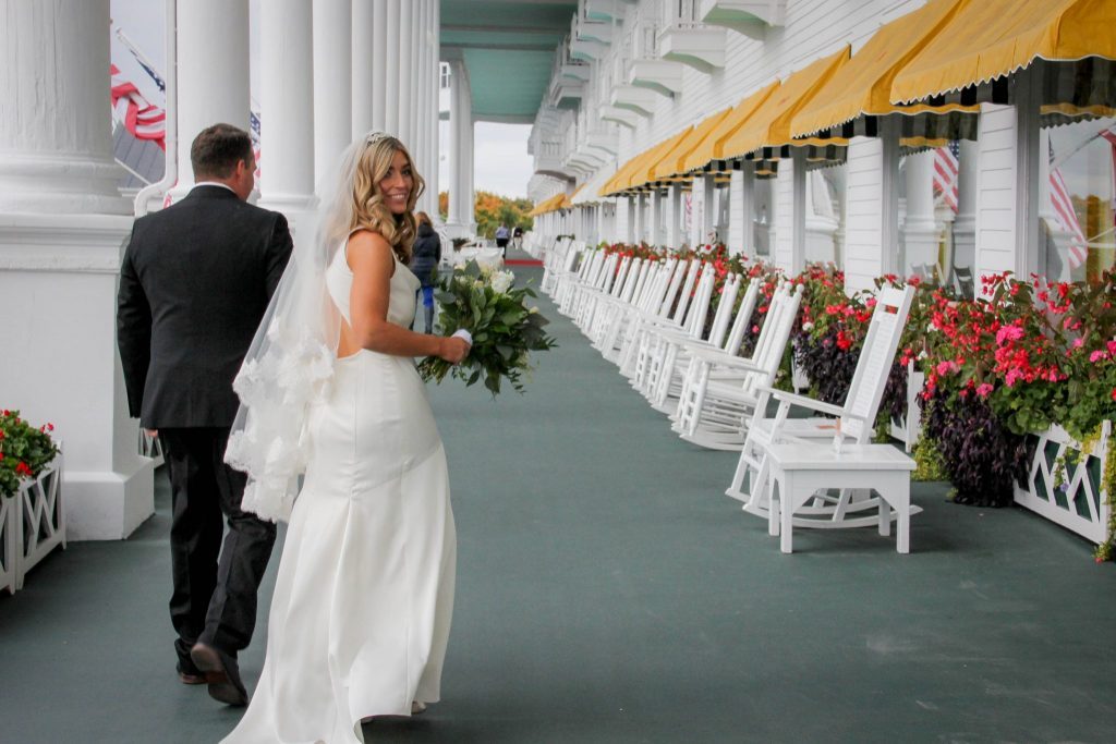 A bride holding a bouquet of flowers turns back to the camera while walking along the front porch of Mackinac Island's Grand Hotel