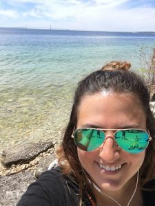 A young woman wearing sunglasses snaps a selfie along the water on Mackinac Island with the Mackinac Bridge in the background