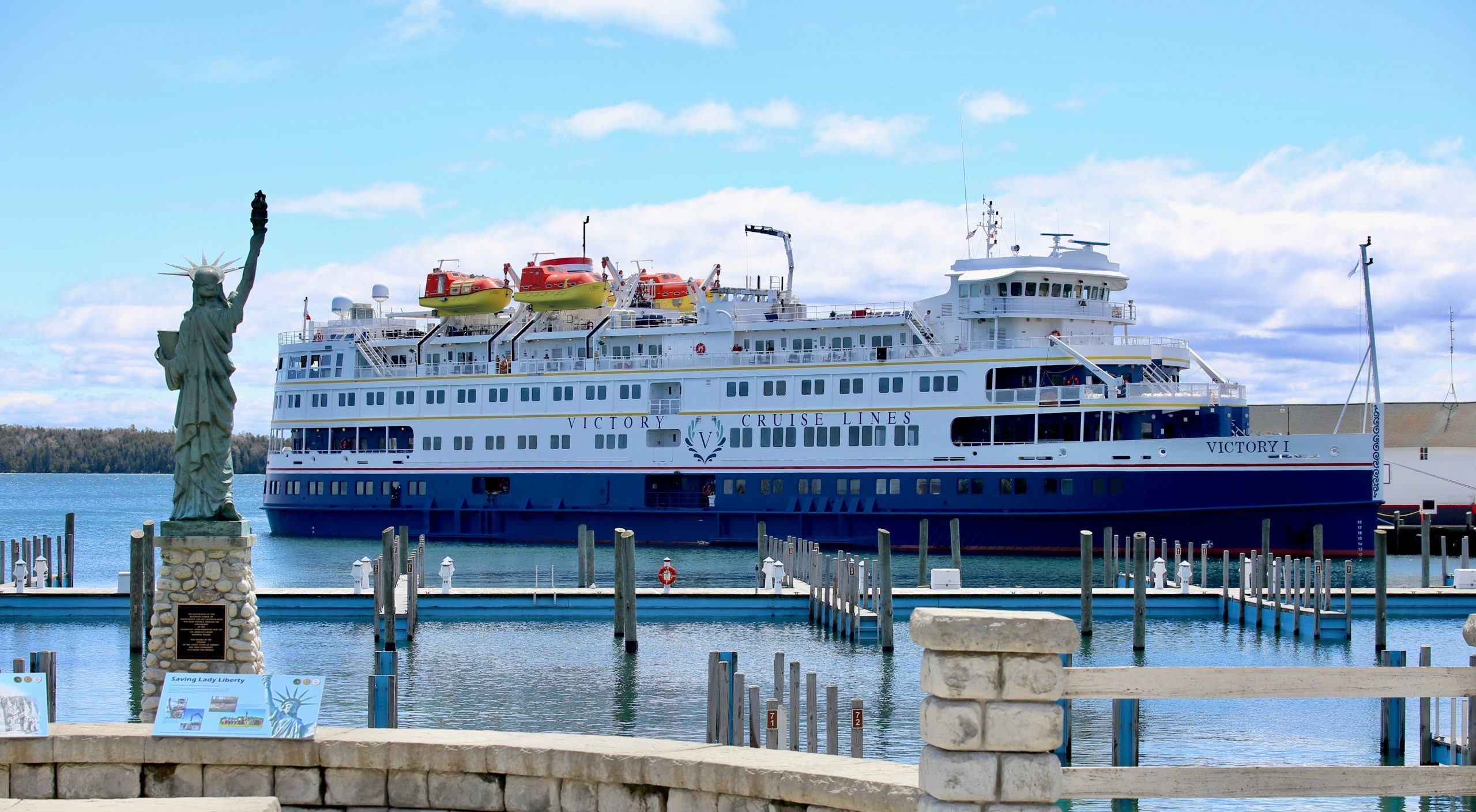 A large Great Lakes cruise ship docks on Mackinac Island with a replica Statue of Liberty in the foreground overlooking the marina