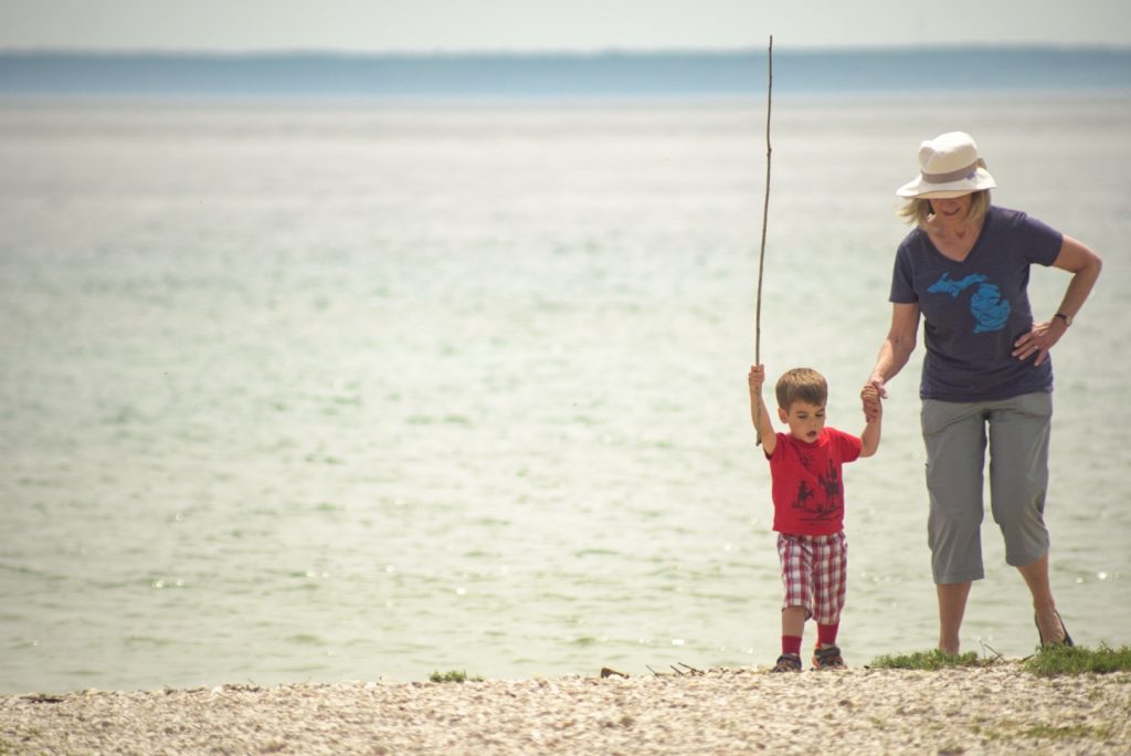 A grandmother holds a young child's hand on a rocky Mackinac Island beach with water behind them