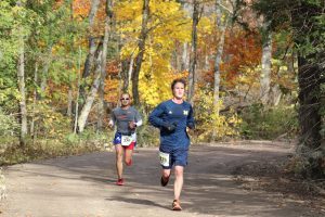 Two runners in a Mackinac Island road race speed downhill through the woods in fall with colored leaves