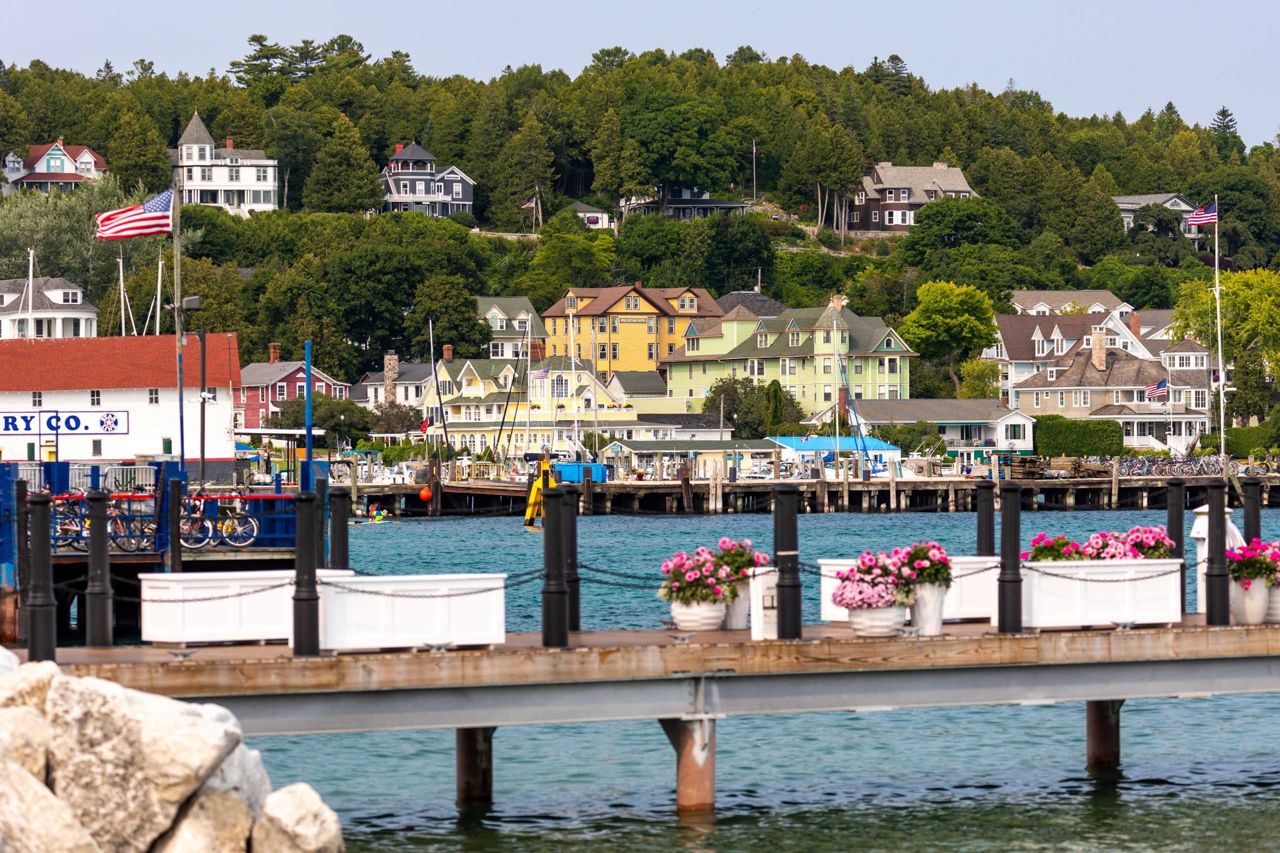 Pots of pink flowers sit on a Mackinac Island dock over the water with hotels and cottages on the bluff in the background