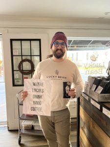 A man inside Mackinac Island’s Lucky Bean Coffeehouse holds a sign that says “Where Locals Drink Good Coffee.”