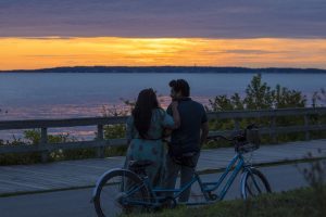 A couple visiting Mackinac Island takes a break from riding a tandem bicycle to watch the sunset along the boardwalk.