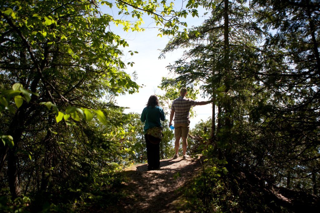 Two people stand at the end of a wooded trail on Mackinac Island and look over the cliff's edge at the water
