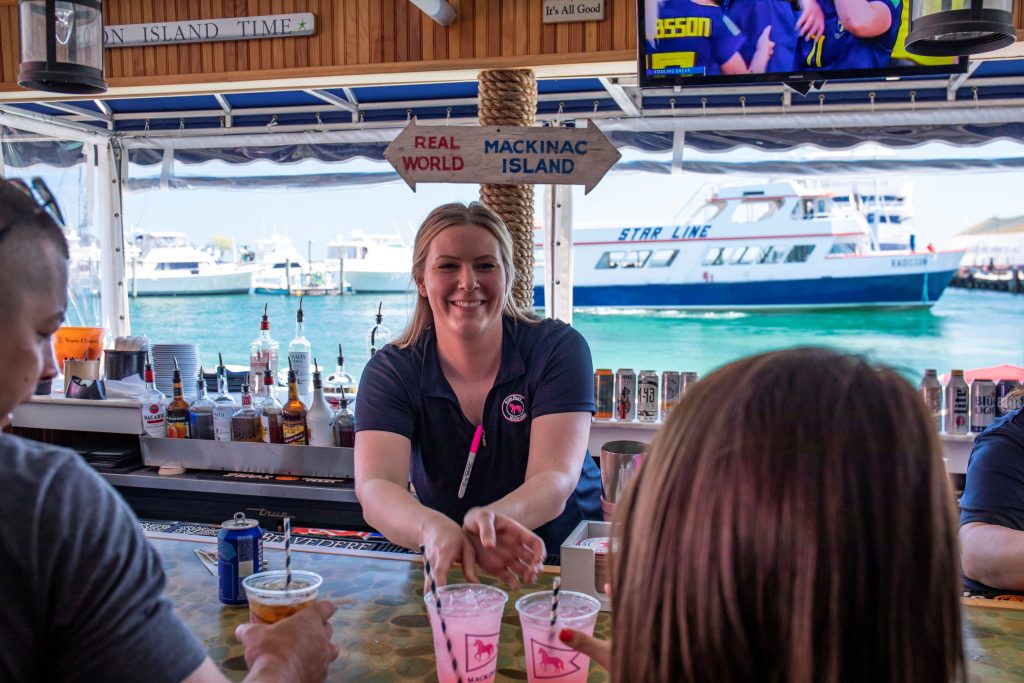 A bartender hands drinks to customers at Pink Pony on Mackinac Island with the water and a ferry boat in the background