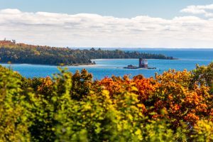 Round Island Lighthouse stands in the blue waters off Mackinac Island with bright fall colors in the foreground on a sunny day