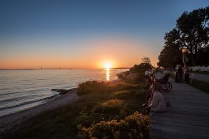 People sit on the Mackinac Island boardwalk and watch the sun set over the water to the west beyond the Mackinac Bridge