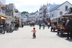 A little boy wearing a bicycle helmet pedals a tiny bike past horses on Main Street in downtown Mackinac Island.