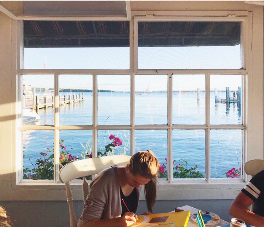A woman paints on canvas in front of a window overlooking the water at Watercolor Café on Mackinac Island