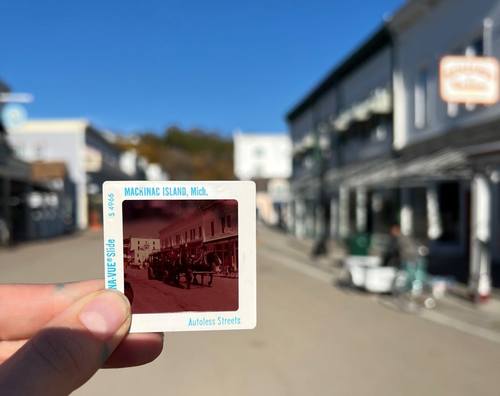 An old photo slide showing a horse-drawn carriage is held in the middle of modern day Mackinac Island's Main Street