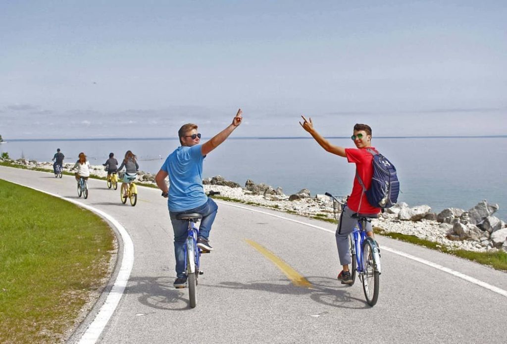 Two guys riding bikes on the road around Mackinac Island look back to the camera and wave