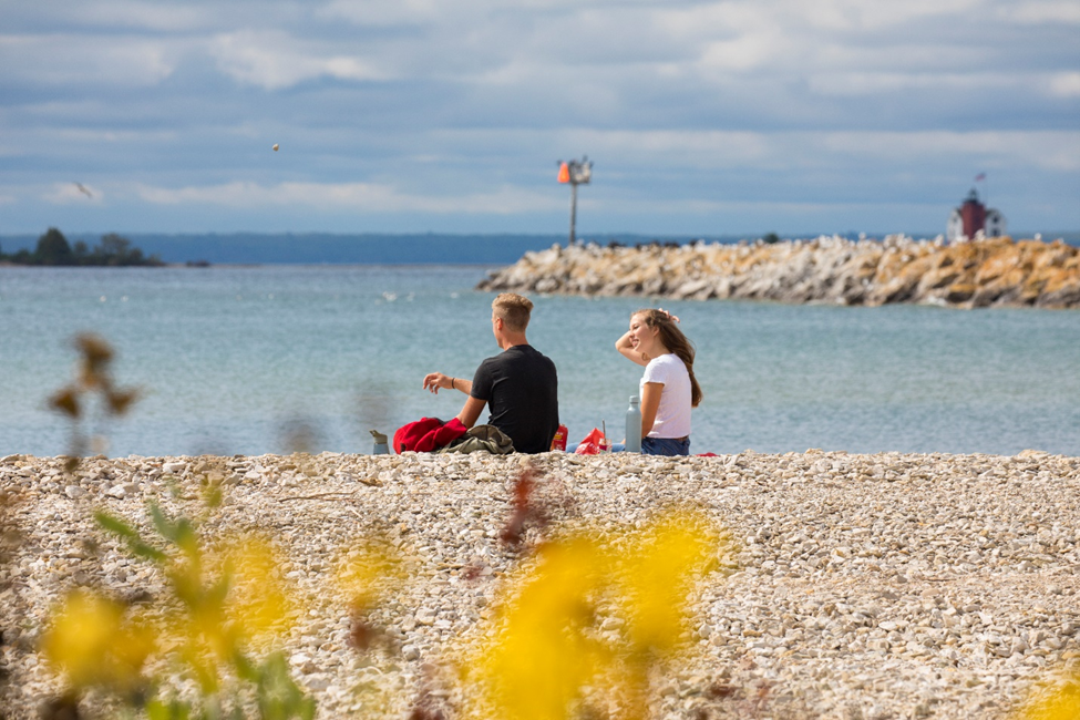 A couple sits on a rocky Mackinac Island beach and has a picnic overlooking the water with flowers in the foreground