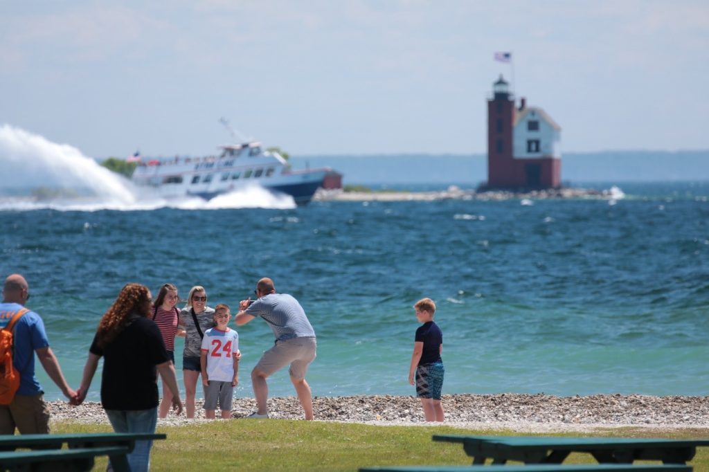 A family takes a photo on a rocky Mackinac Island beach with a ferry boat and Round Island Lighthouse in the background.