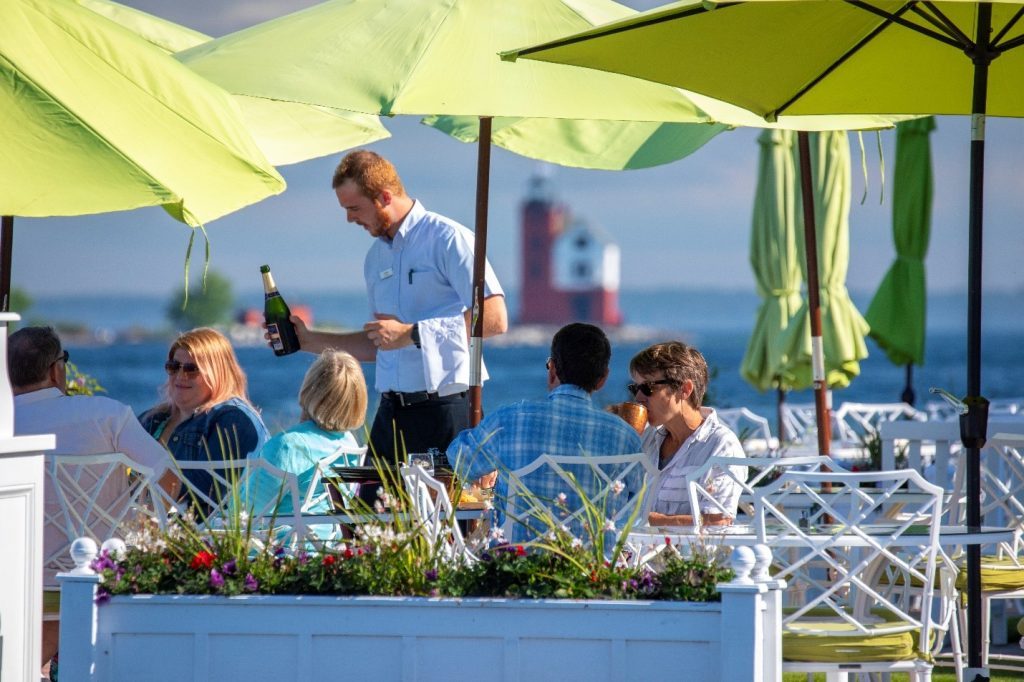 A waiter pours a bottle of champagne for diners on a Mackinac Island restaurant patio with a lighthouse in the background. 
