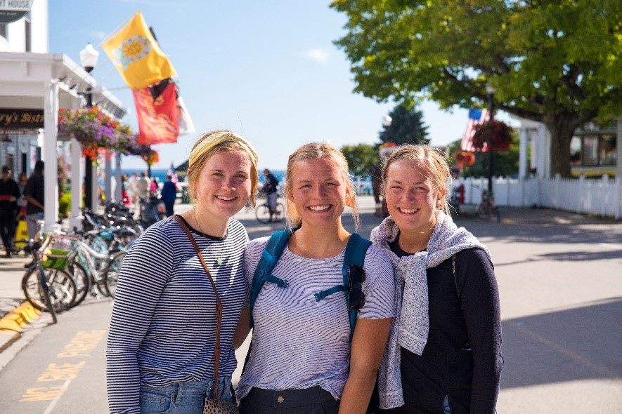 Three young women pose for a photo on Mackinac Island’s Main Street on a sunny day with water in the background.