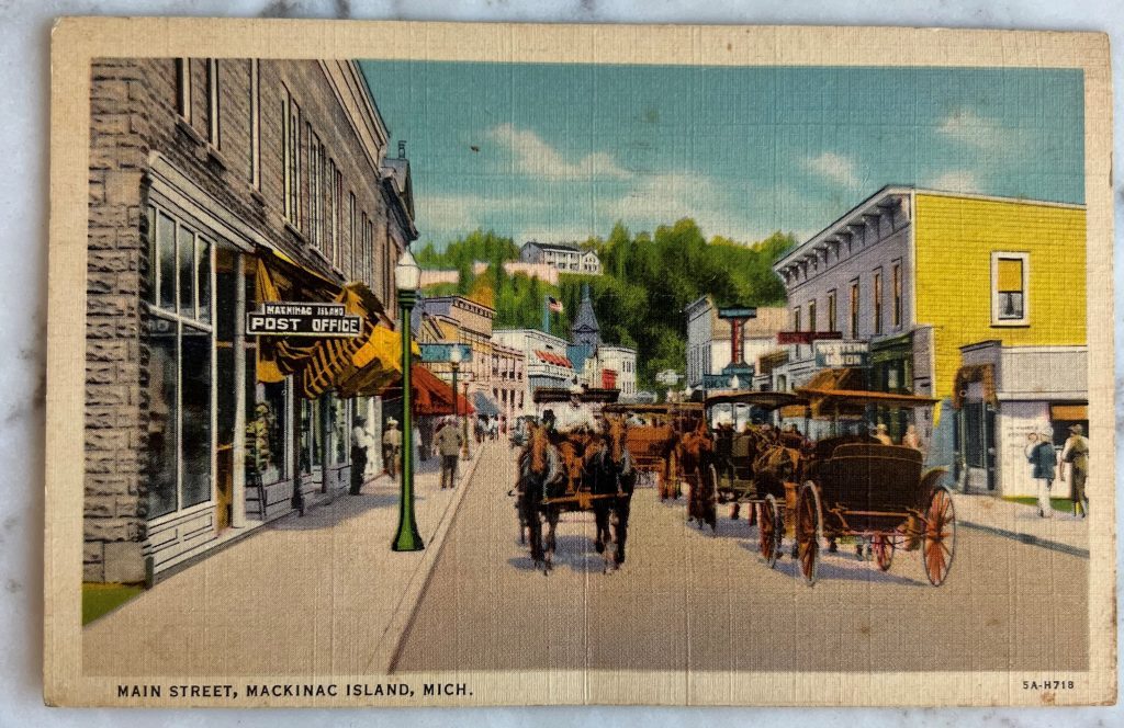 An old postcard showing horse-drawn carriages go past the Mackinac Island Post Office on Main Street