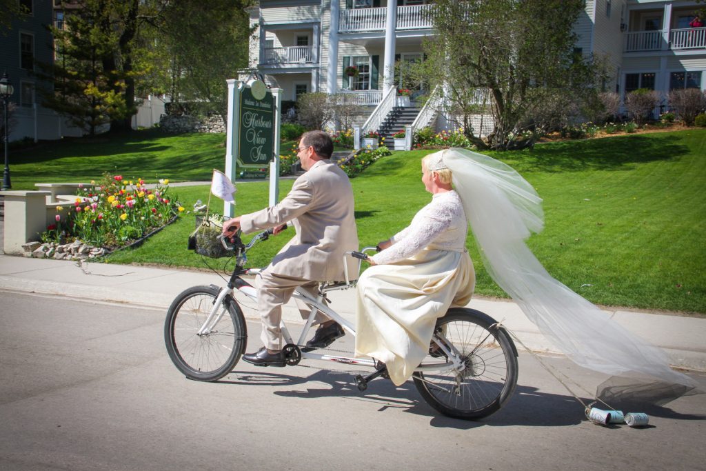 A bride and groom ride a tandem bicycle past Mackinac Island's Harbour View Inn, with cans clinking on the street behind the back tire