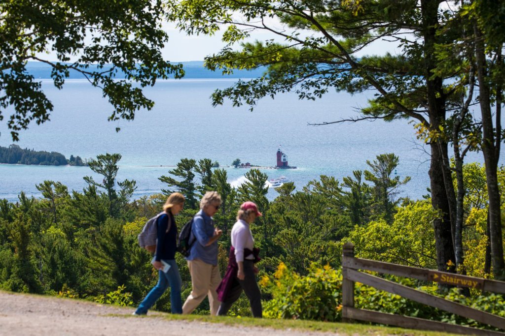 A group of three women walks through the interior of Mackinac Island with water, a ferry boat and Round Island Lighthouse in the background