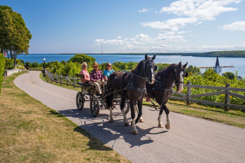 A horse drawn carriage takes Mackinac Island visitors up a hill with a stunning view of blue waters in the background