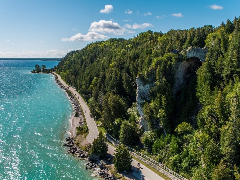 Arch Rock looms high above Mackinac Island’s M-185 roadway in an aerial drone shot of the forested Lake Huron coastline.