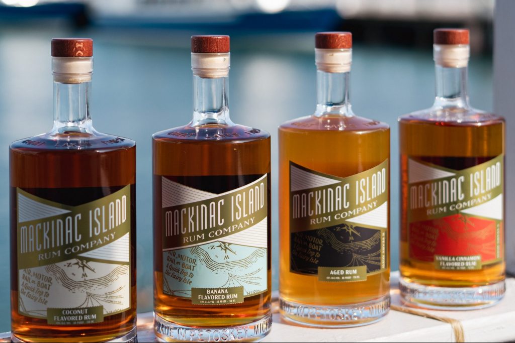Four bottles of Mackinac Island Rum Company liquor sit on a waterfront ledge with a ferry boat in the background.