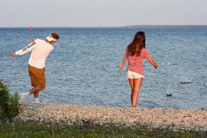 A young man and a young woman skips stones into the refreshing blue water at a rocky Mackinac Island beach
