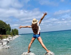 A young woman stands with arms wide apart on two rocks at the edge of the water on Mackinac Island
