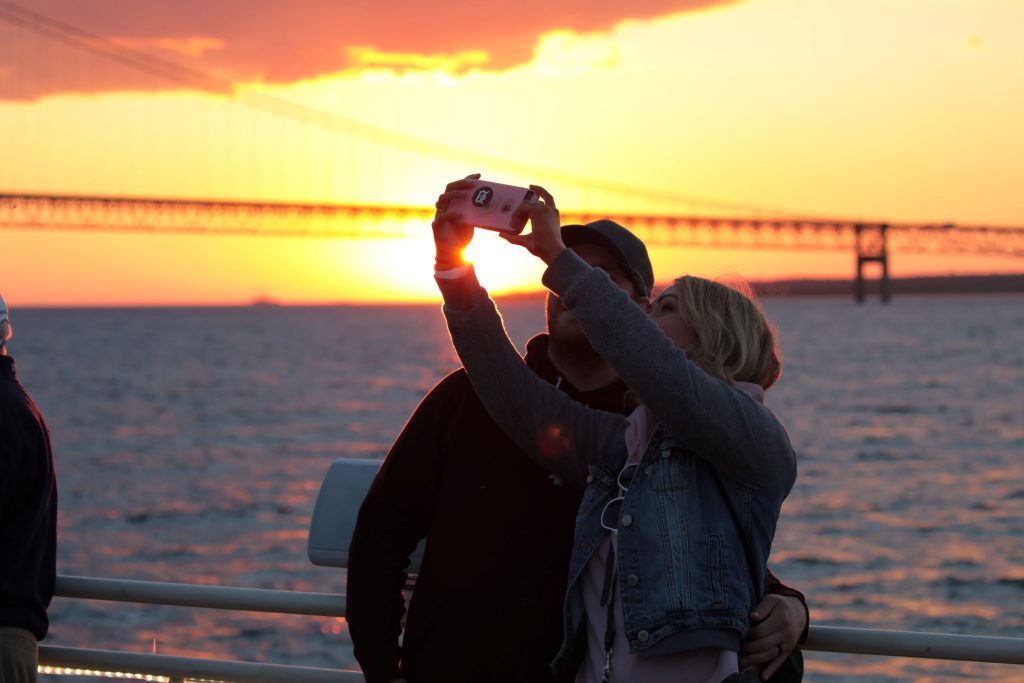 A couple snaps a selfie on a Mackinac Island Sip ‘n Sail Cruise as the boat nears the Mackinac Bridge at sunset