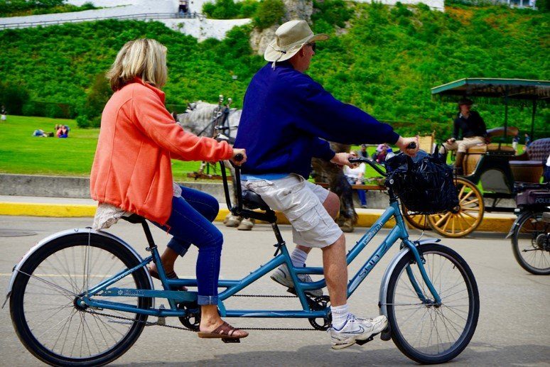 Couple Biking Past Carriage – Mackinac Island Tourism Bureau