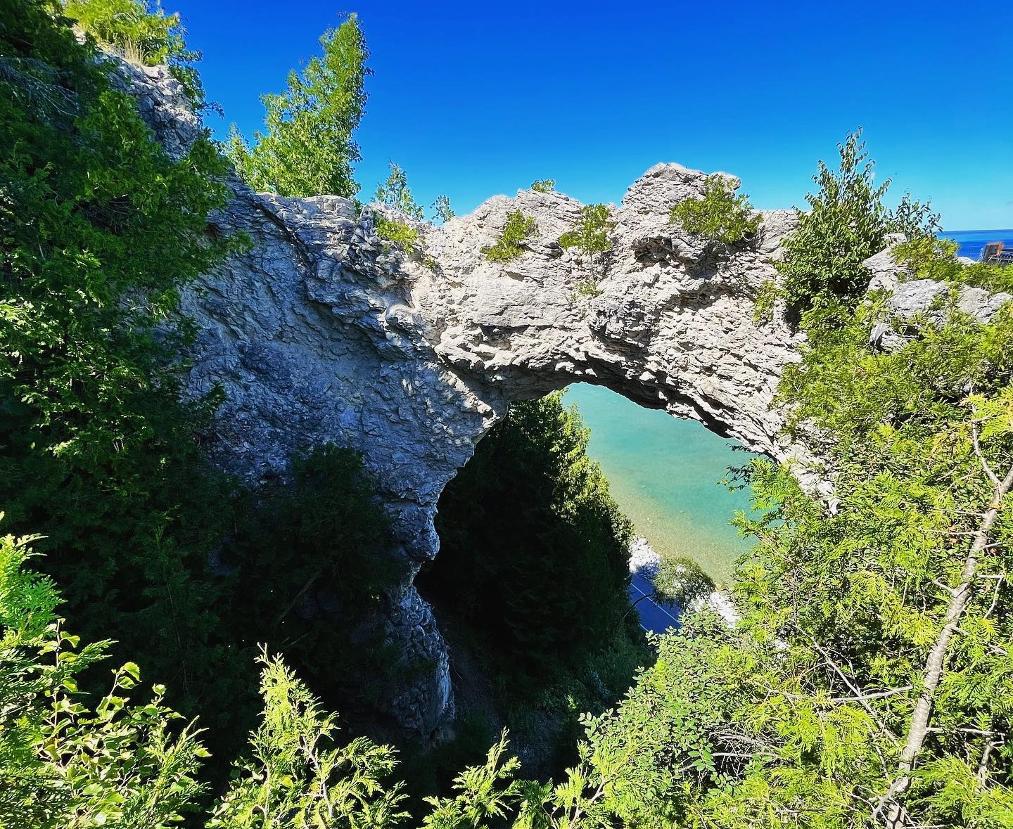 Mackinac Island’s Arch Rock frames the road and water below with bright blue sky above and green trees